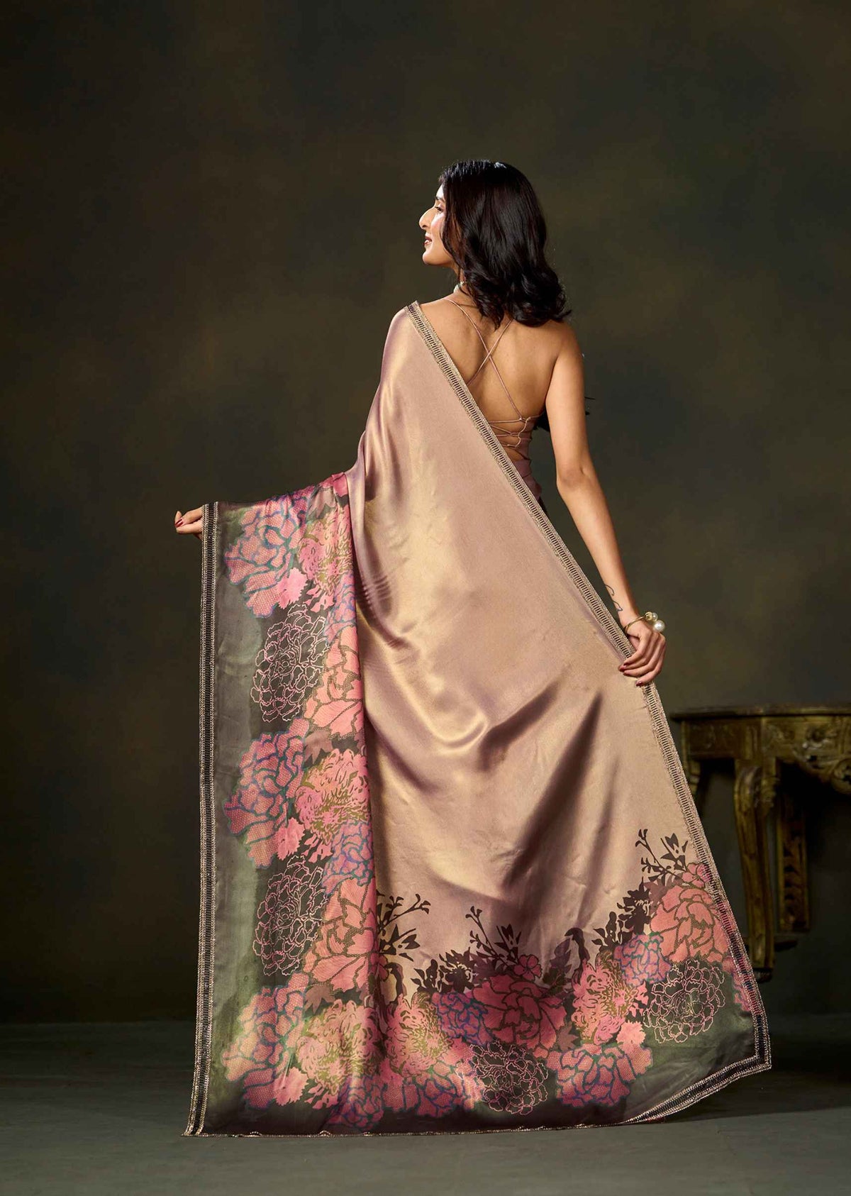 A woman is shown from behind wearing a draped beige saree with a wide border featuring large pink and purple floral patterns, posed against a dark studio background.