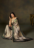 A woman wearing a flowing satin saree with a black, green, and gold floral print, seated in a studio setting with a dark textured background and a small wooden table on the right.