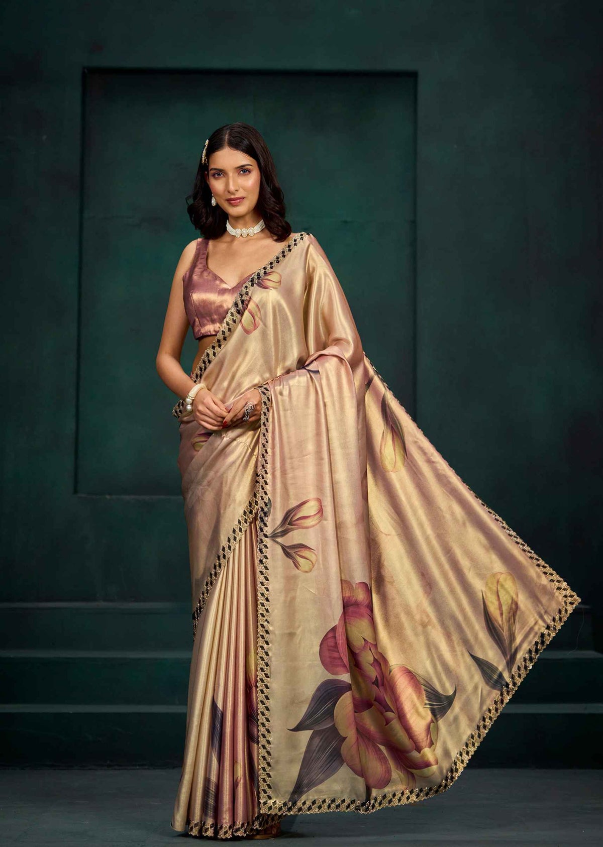 A woman models a beige silk saree with a metallic pink blouse. The saree features a large floral print in pink, yellow, and green, with a black and gold border.