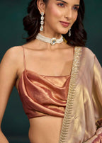 A close-up of a woman wearing a metallic rose-gold satin blouse with thin straps and a draped champagne-gold saree with a gold embroidered border, paired with a white choker necklace and dangling earrings, against a dark green background.