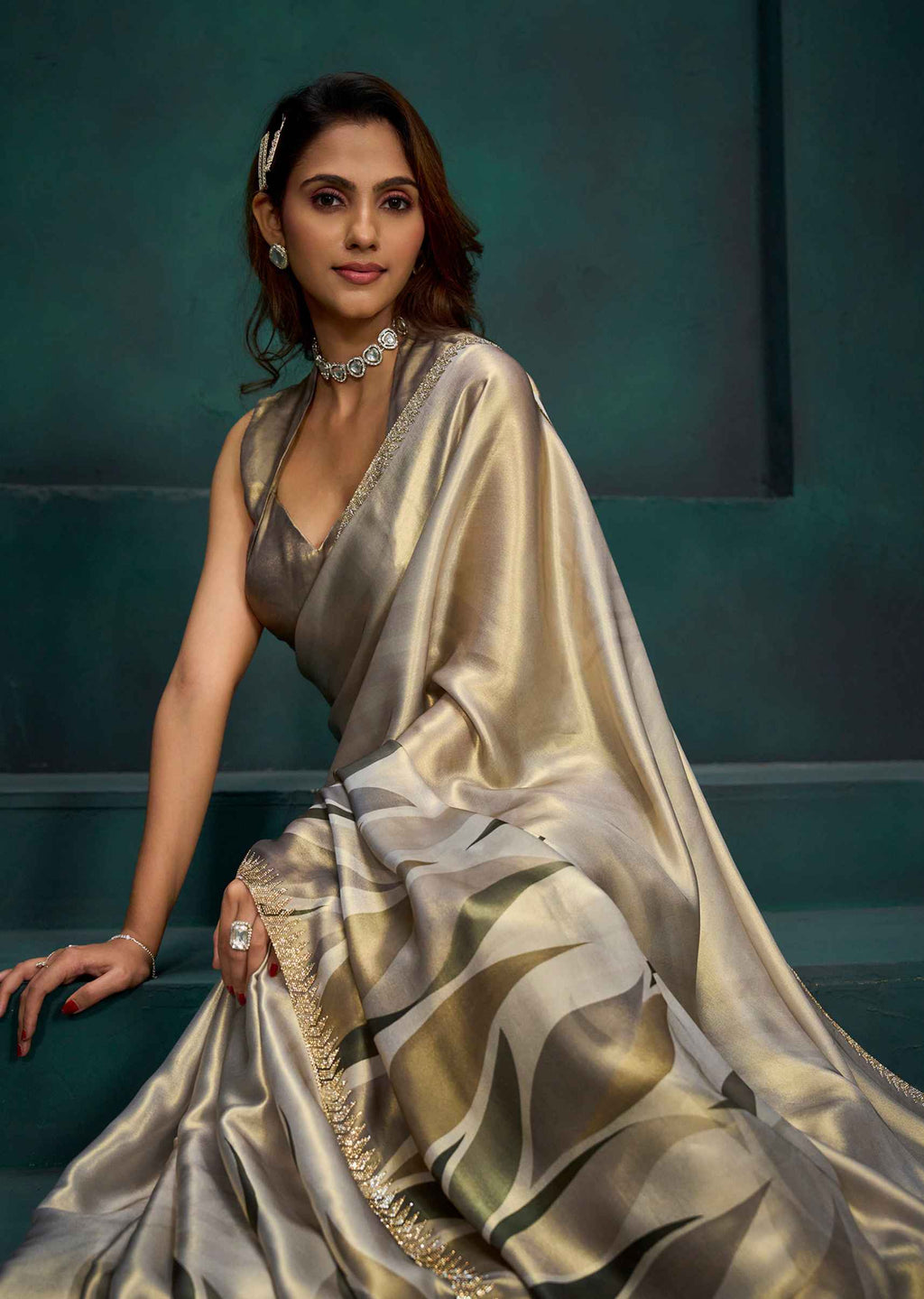 A woman wearing a shimmering champagne-gold saree with a matching blouse, seated against a dark teal studio backdrop. The saree has a satin sheen with wavy metallic stripes and a gold embroidered border.