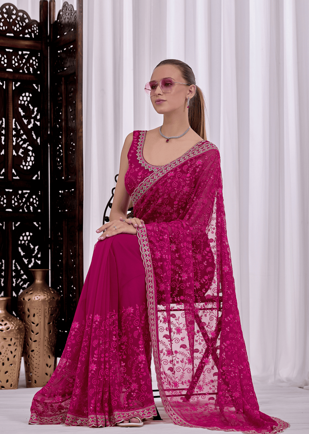 A woman models a bright magenta pink embroidered saree with a matching blouse, standing in front of white curtains and a dark wooden screen.