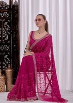 A woman models a bright magenta pink embroidered saree with a matching blouse, standing in front of white curtains and a dark wooden screen.