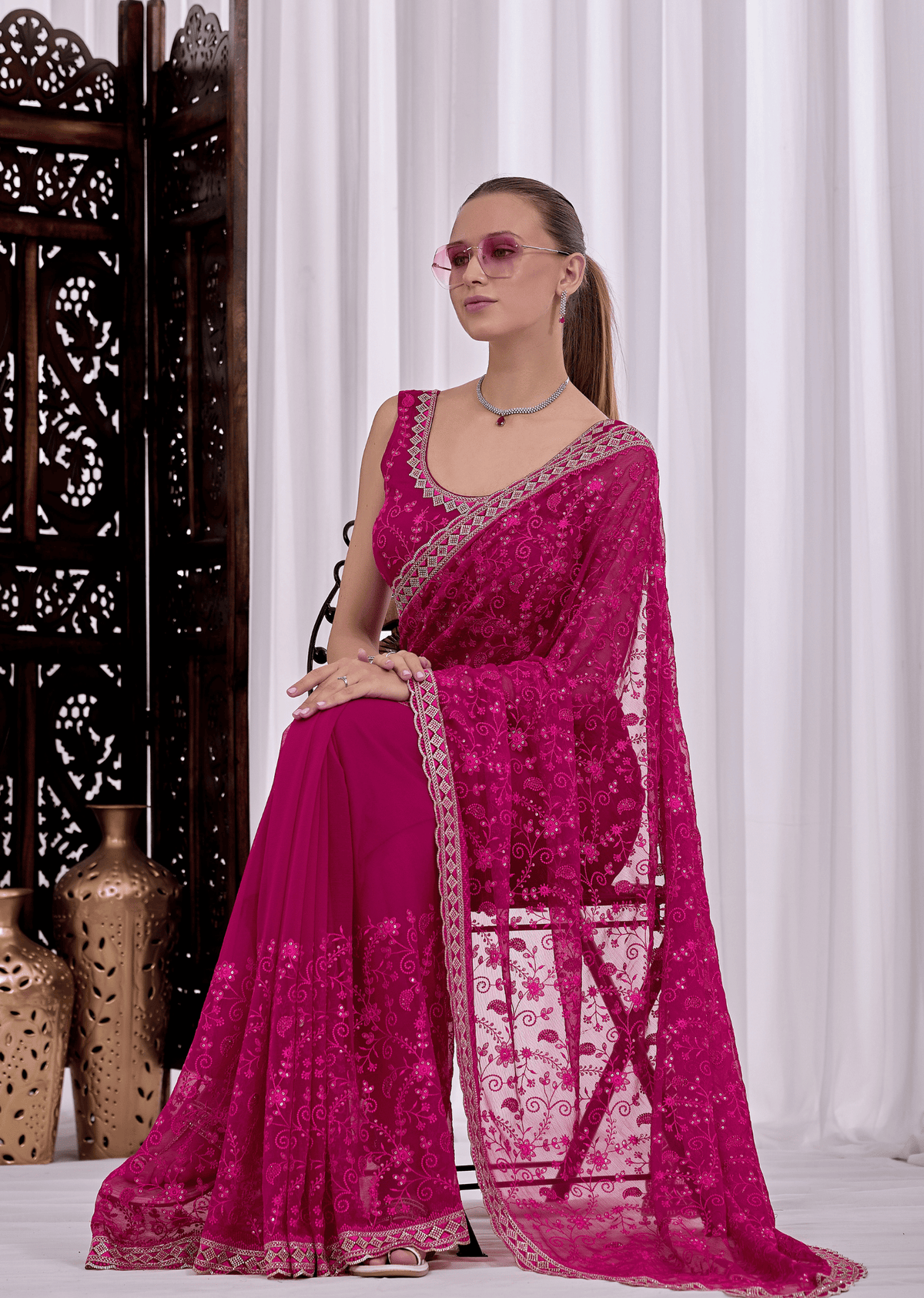 A woman models a bright magenta pink embroidered saree with a matching blouse, standing in front of white curtains and a dark wooden screen.