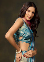 A woman modeling a blue patterned sleeveless blouse with a side cutout and matching draped sari, posed against a dark brown studio background.