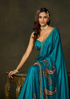 A woman wearing a teal blue saree with gold and brown paisley embroidery, seated on an ornate wooden table against a dark brown studio background.
