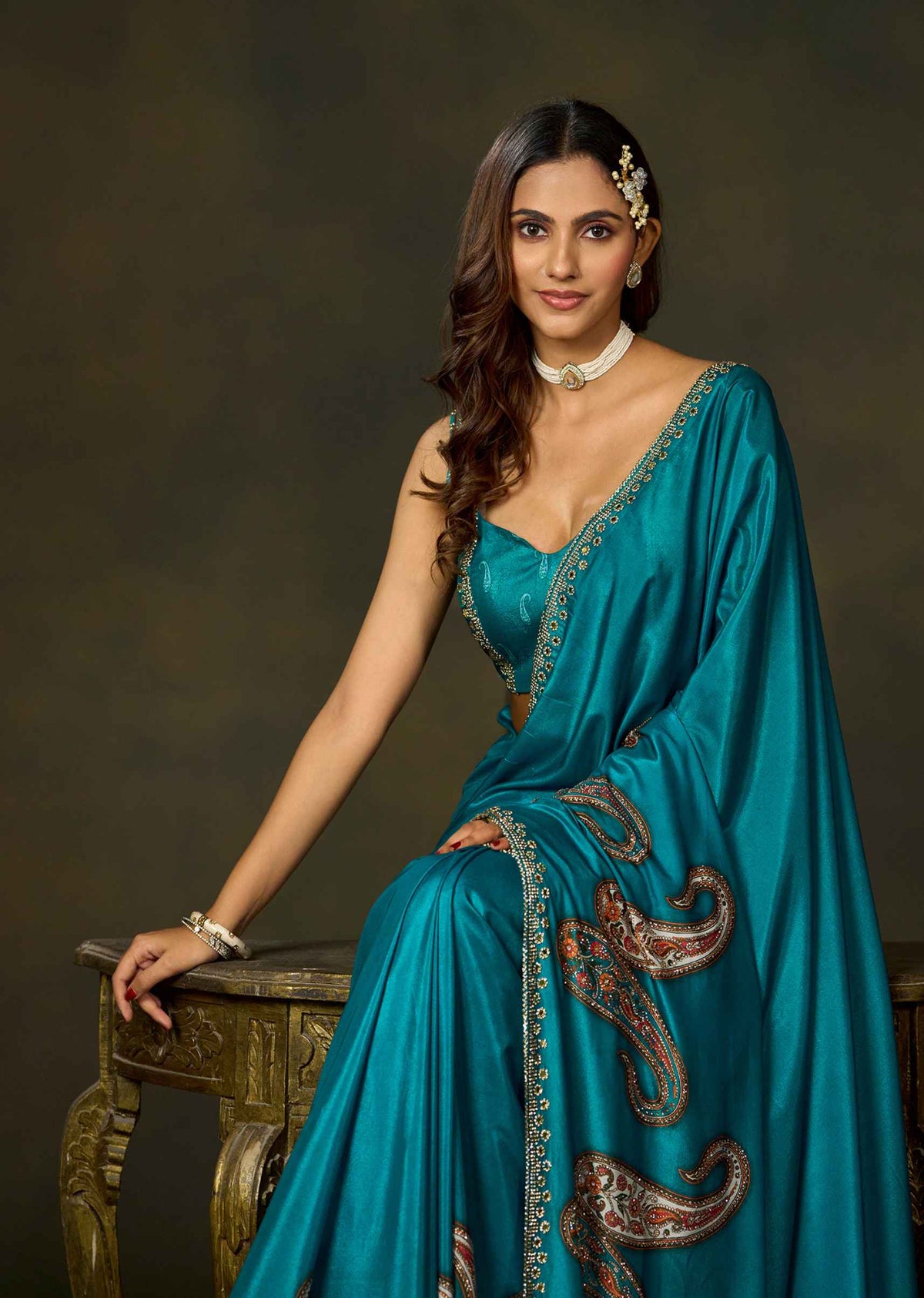 A woman wearing a teal blue saree with gold and brown paisley embroidery, seated on an ornate wooden table against a dark brown studio background.