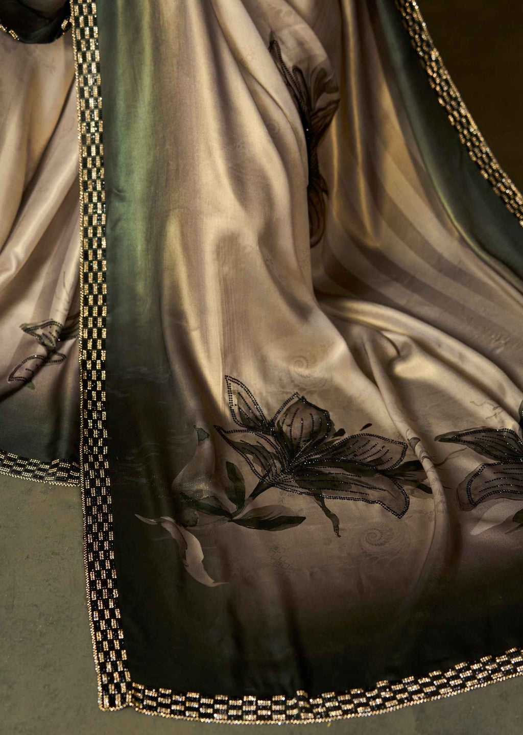 A close-up of a draped satin fabric with a dark olive green to taupe gradient, featuring black floral illustrations and a decorative black-and-gold checkered border.