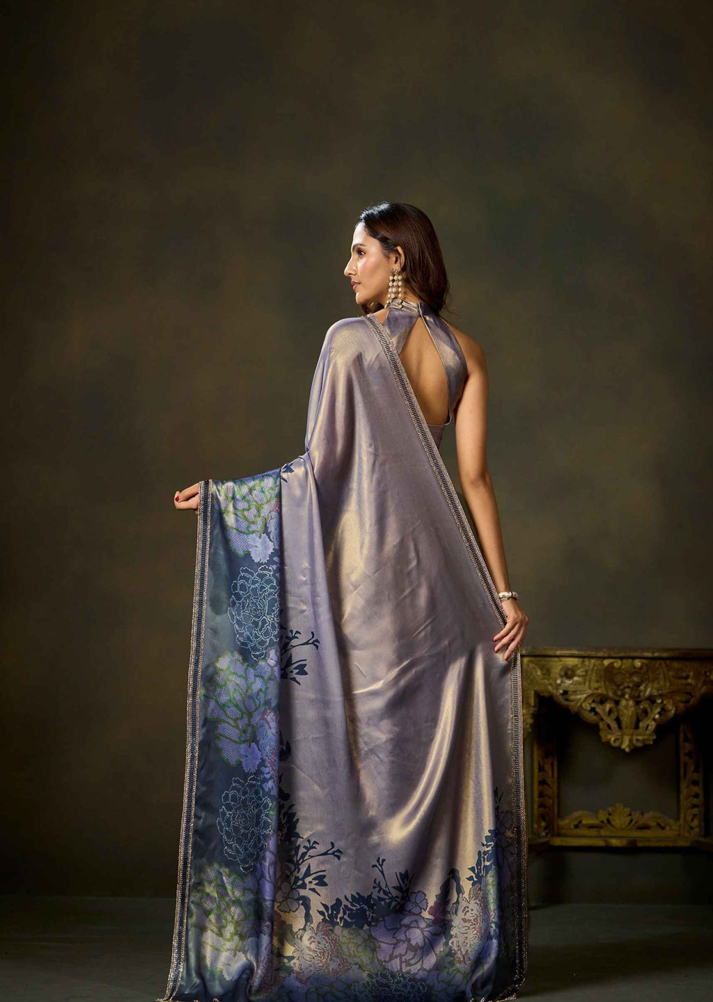A woman wearing a draped lavender-purple saree with blue floral patterns, shown from the back against a dark brown studio backdrop.