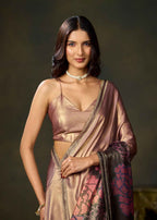 A woman modeling a metallic champagne pink saree with a matching sleeveless blouse, featuring a dark border with pink floral embroidery and a pearl choker, set against a dark brown studio background.