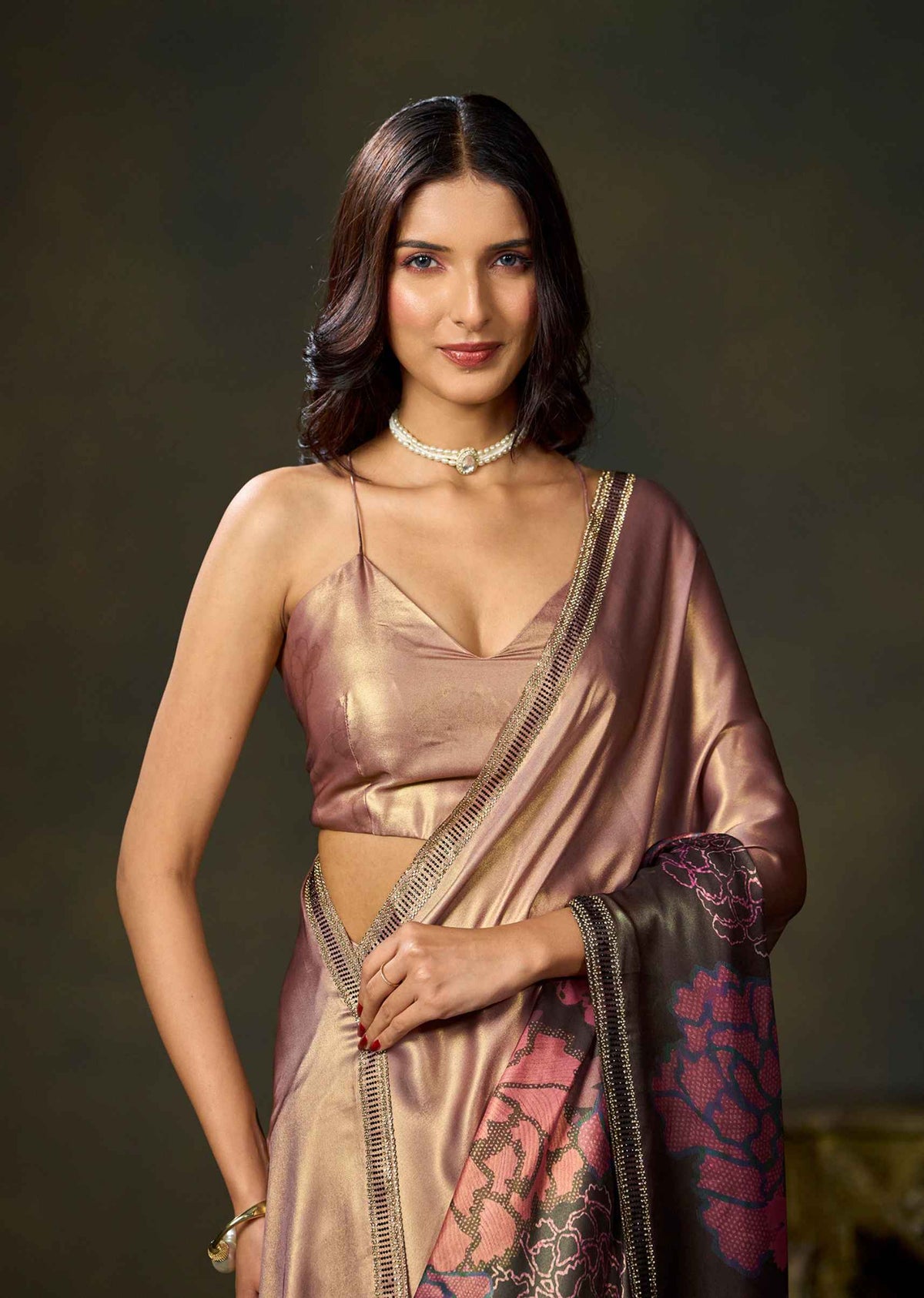 A woman modeling a metallic champagne pink saree with a matching sleeveless blouse, featuring a dark border with pink floral embroidery and a pearl choker, set against a dark brown studio background.