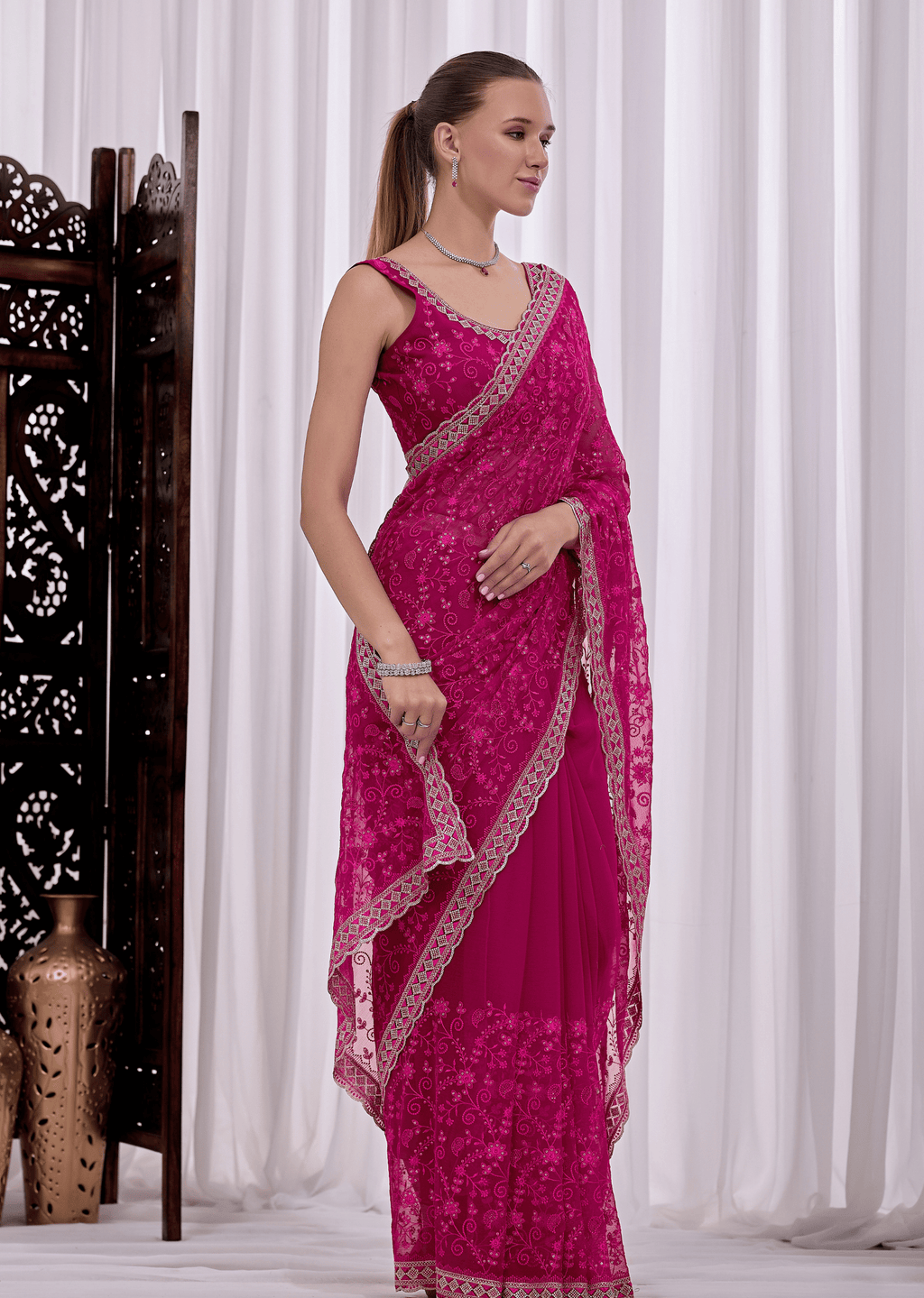 A woman modeling a bright magenta pink embroidered saree with a matching sleeveless blouse, standing in front of white draped curtains.