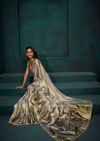 A woman wearing a flowing metallic champagne-gold gown with a draped cape-style overlay, seated on dark teal steps against a matching dark teal wall.