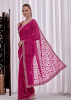 A woman modeling a bright fuchsia pink embroidered saree with a matching sheer dupatta, set against a white curtain backdrop.