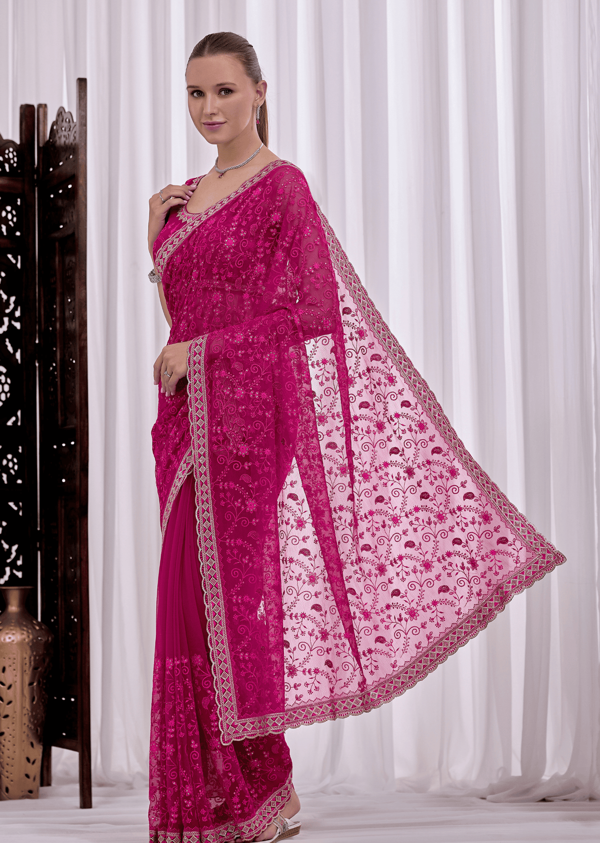 A woman modeling a bright fuchsia pink embroidered saree with a matching sheer dupatta, set against a white curtain backdrop.