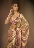 A woman modeling a metallic champagne-gold saree with pink floral patterns and a matching pink blouse, styled with a pearl necklace and bracelet against a warm brown studio background.
