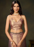 A woman modeling a metallic rose gold two-piece outfit consisting of a spaghetti-strap crop top and a matching skirt, styled with a pearl choker and bracelet against a dark brown studio background.