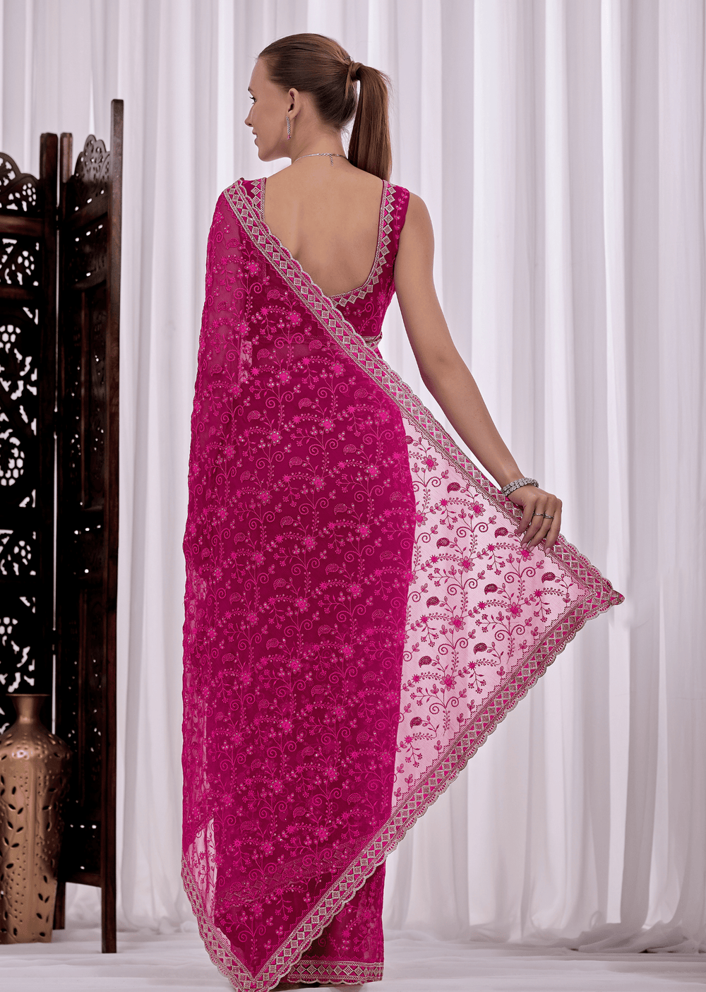 A woman modeling a bright pink embroidered saree with a matching blouse, shown from the back in a studio setting with white curtains and a decorative screen.