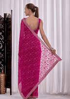 A woman modeling a bright pink embroidered saree with a matching blouse, shown from the back in a studio setting with white curtains and a decorative screen.