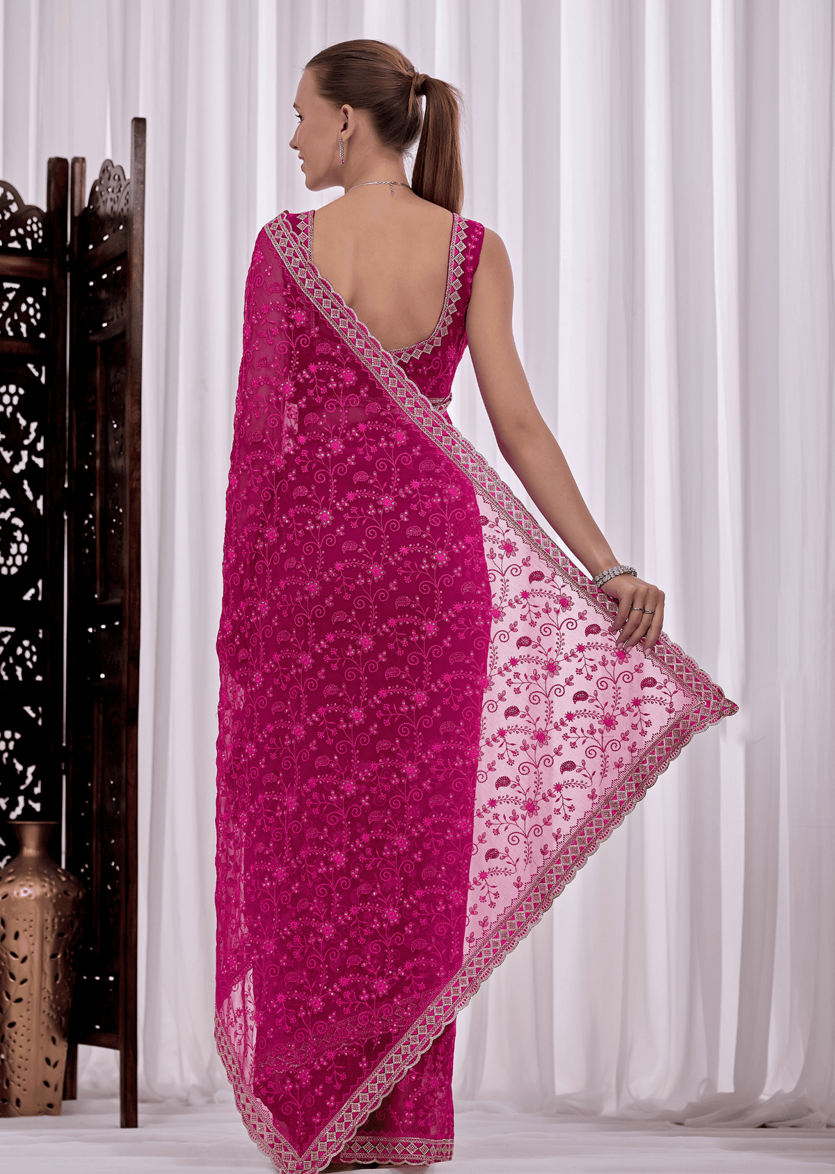 A woman modeling a bright pink embroidered saree with a matching blouse, shown from the back in a studio setting with white curtains and a decorative screen.