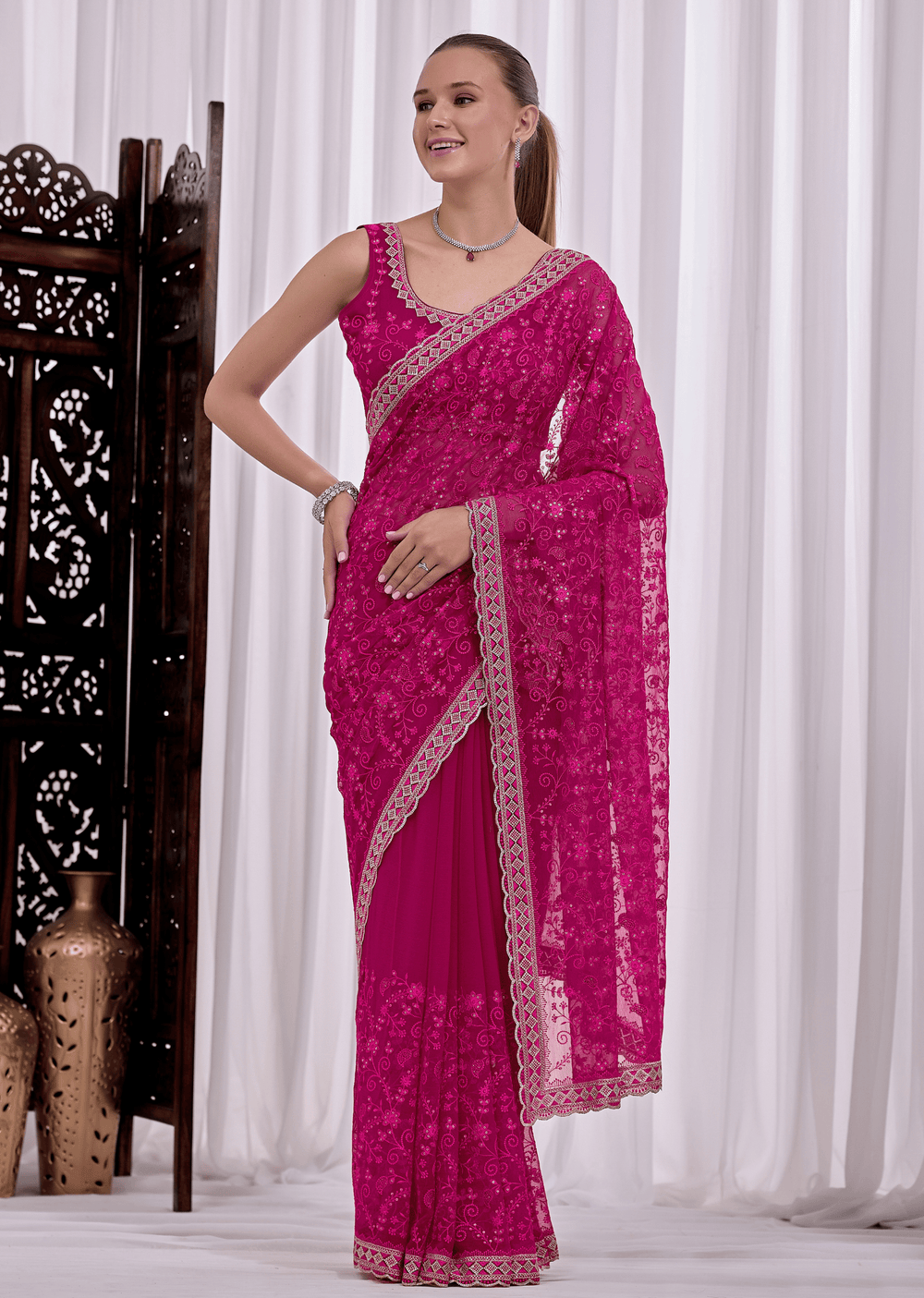 A woman modeling a bright magenta pink saree with intricate silver floral embroidery and a matching embroidered border, posed in front of white curtains.