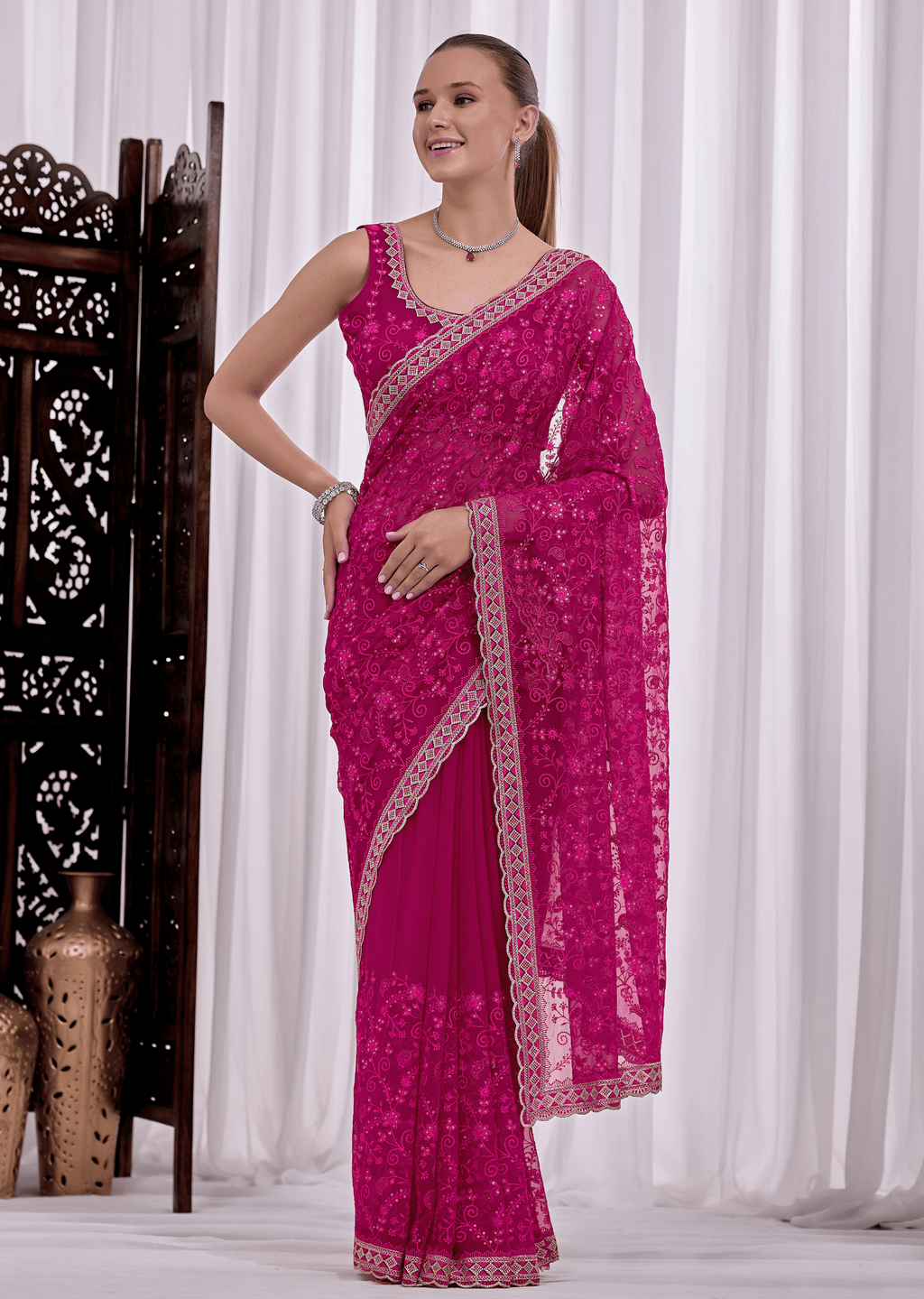 A woman modeling a bright magenta pink saree with intricate silver floral embroidery and a matching embroidered border, posed in front of white curtains.