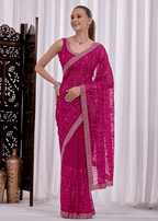 A woman modeling a bright magenta pink saree with intricate silver floral embroidery and a matching embroidered border, posed in front of white curtains.