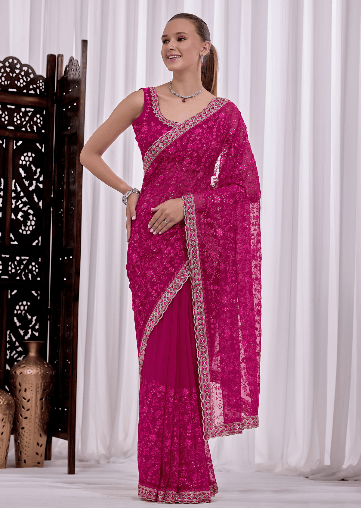 A woman modeling a bright magenta pink saree with intricate silver floral embroidery and a matching embroidered border, posed in front of white curtains.