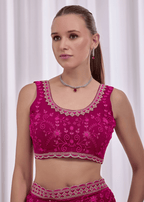 A woman modeling a bright magenta pink embroidered Indian ethnic blouse with intricate silver mirror work and floral patterns, paired with matching pink bottoms against a white curtain background.