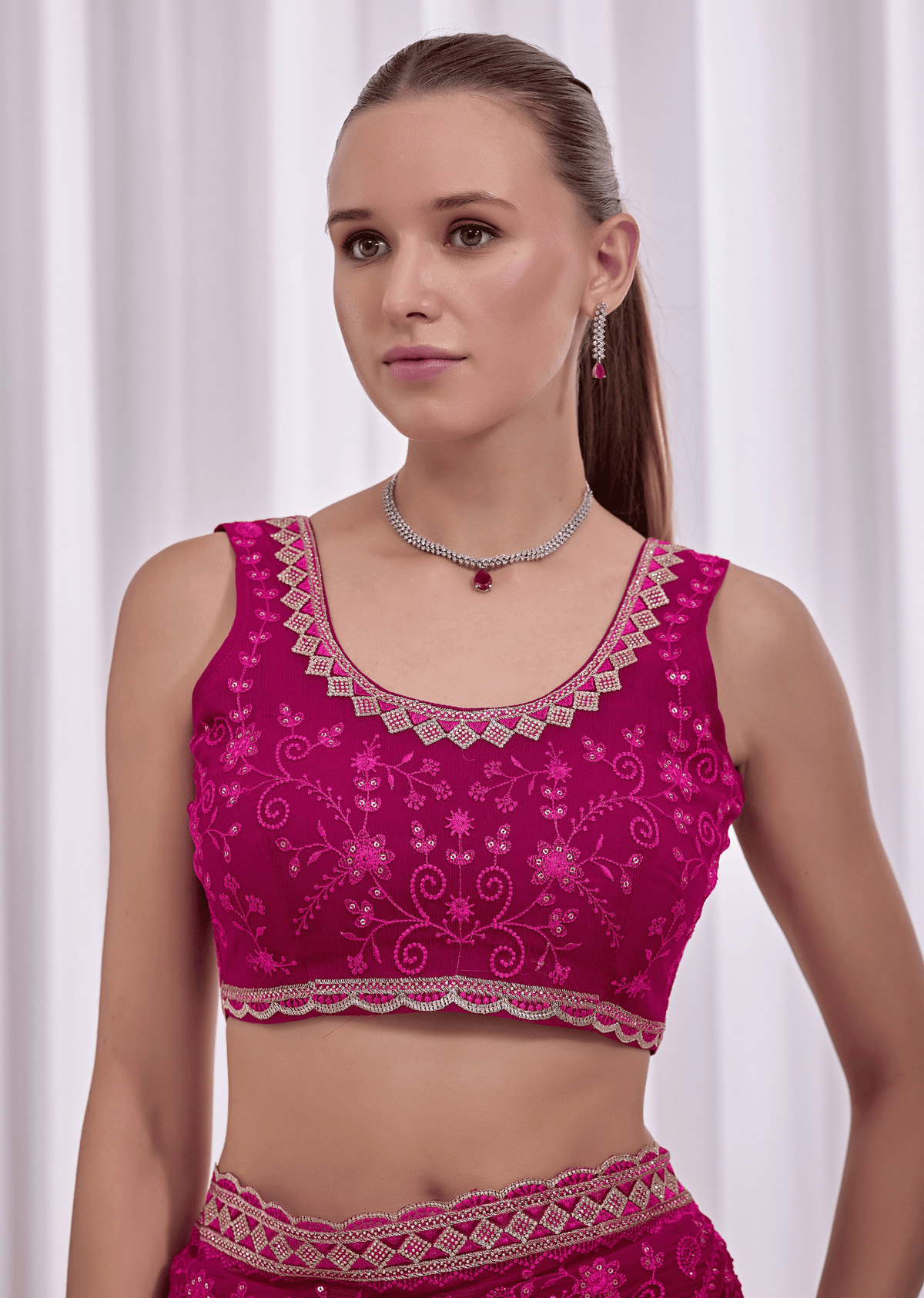 A woman modeling a bright magenta pink embroidered Indian ethnic blouse with intricate silver mirror work and floral patterns, paired with matching pink bottoms against a white curtain background.