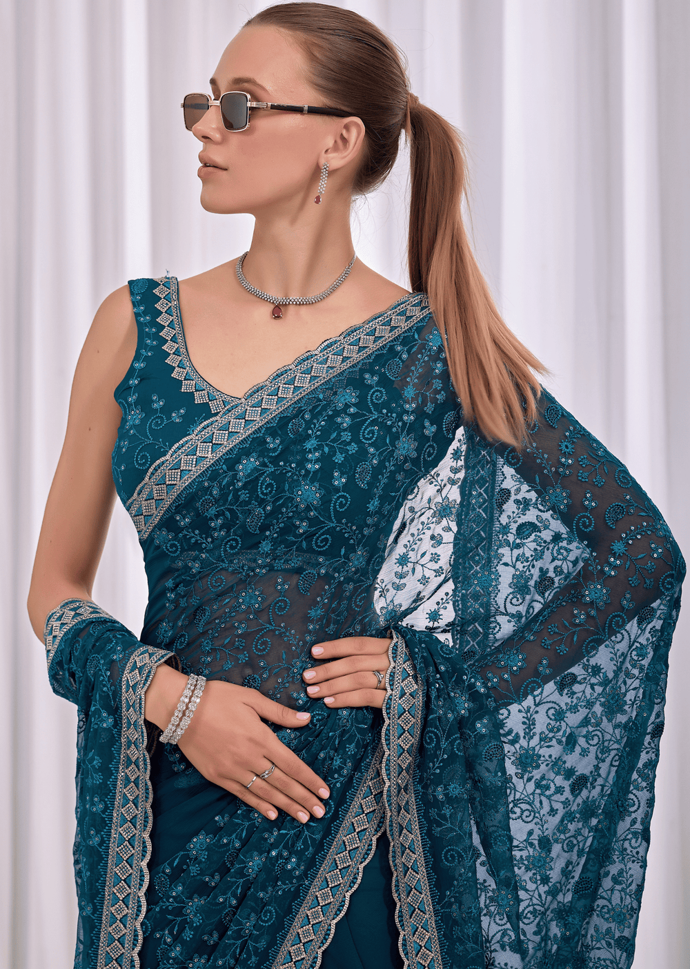 A woman wearing a teal blue embroidered saree with gold borders, a matching blouse, and a dupatta, styled with sunglasses, a necklace, and bracelets against a white curtain background.