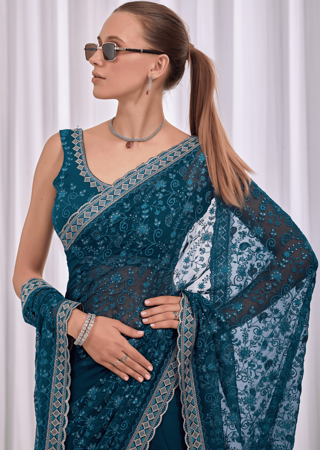 A woman wearing a teal blue embroidered saree with gold borders, a matching blouse, and a dupatta, styled with sunglasses, a necklace, and bracelets against a white curtain background.
