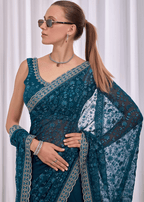 A woman wearing a teal blue embroidered saree with gold borders, a matching blouse, and a dupatta, styled with sunglasses, a necklace, and bracelets against a white curtain background.