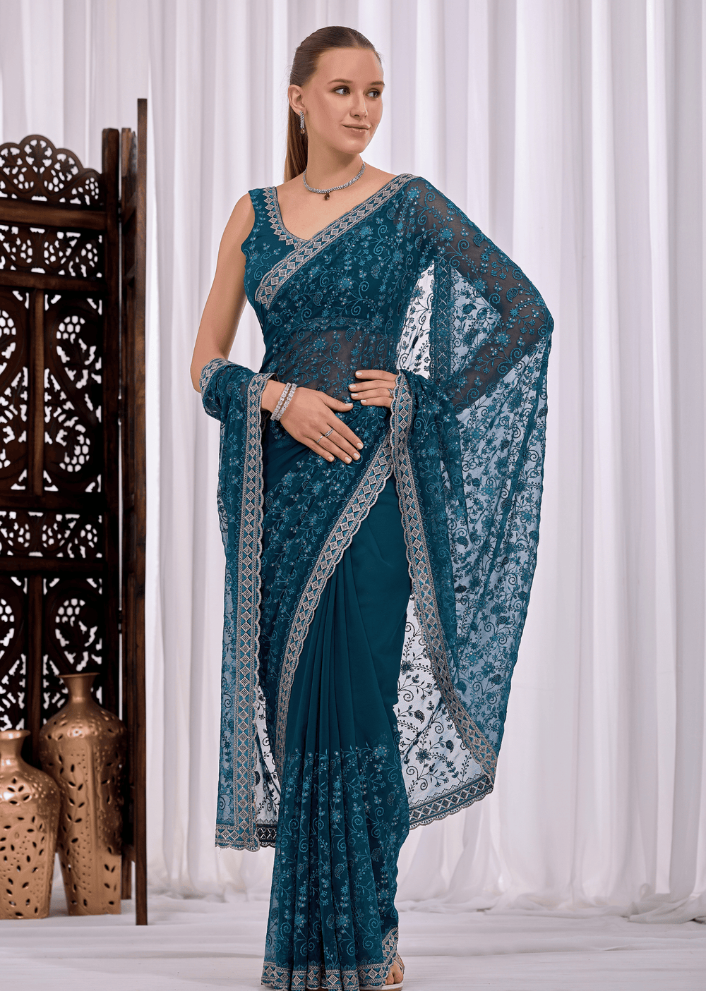 A woman modeling a teal blue saree with intricate silver floral embroidery and a matching embroidered border, standing in front of a white curtain backdrop.