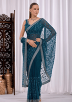 A woman modeling a teal blue saree with intricate silver floral embroidery and a matching embroidered border, standing in front of a white curtain backdrop.