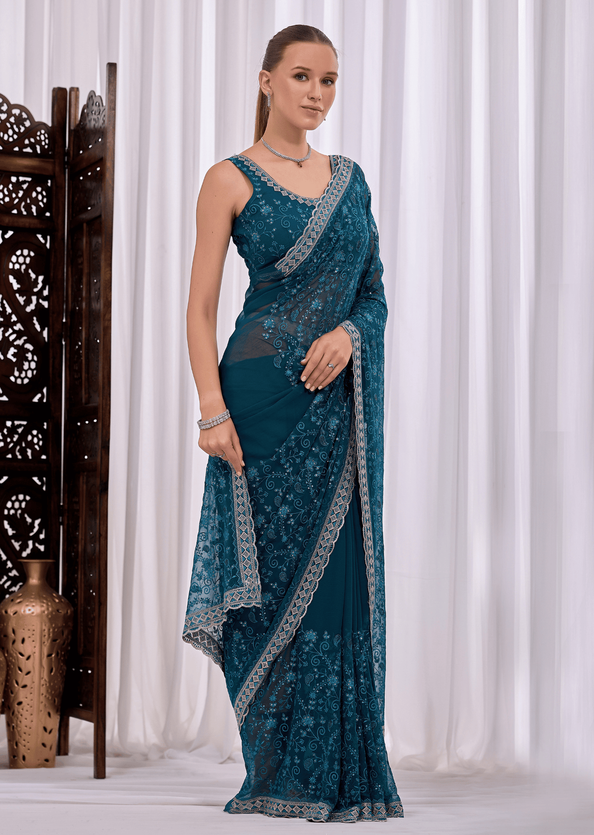 A woman modeling a teal blue saree with intricate silver floral embroidery and a matching embroidered border, posed against a white curtain backdrop.