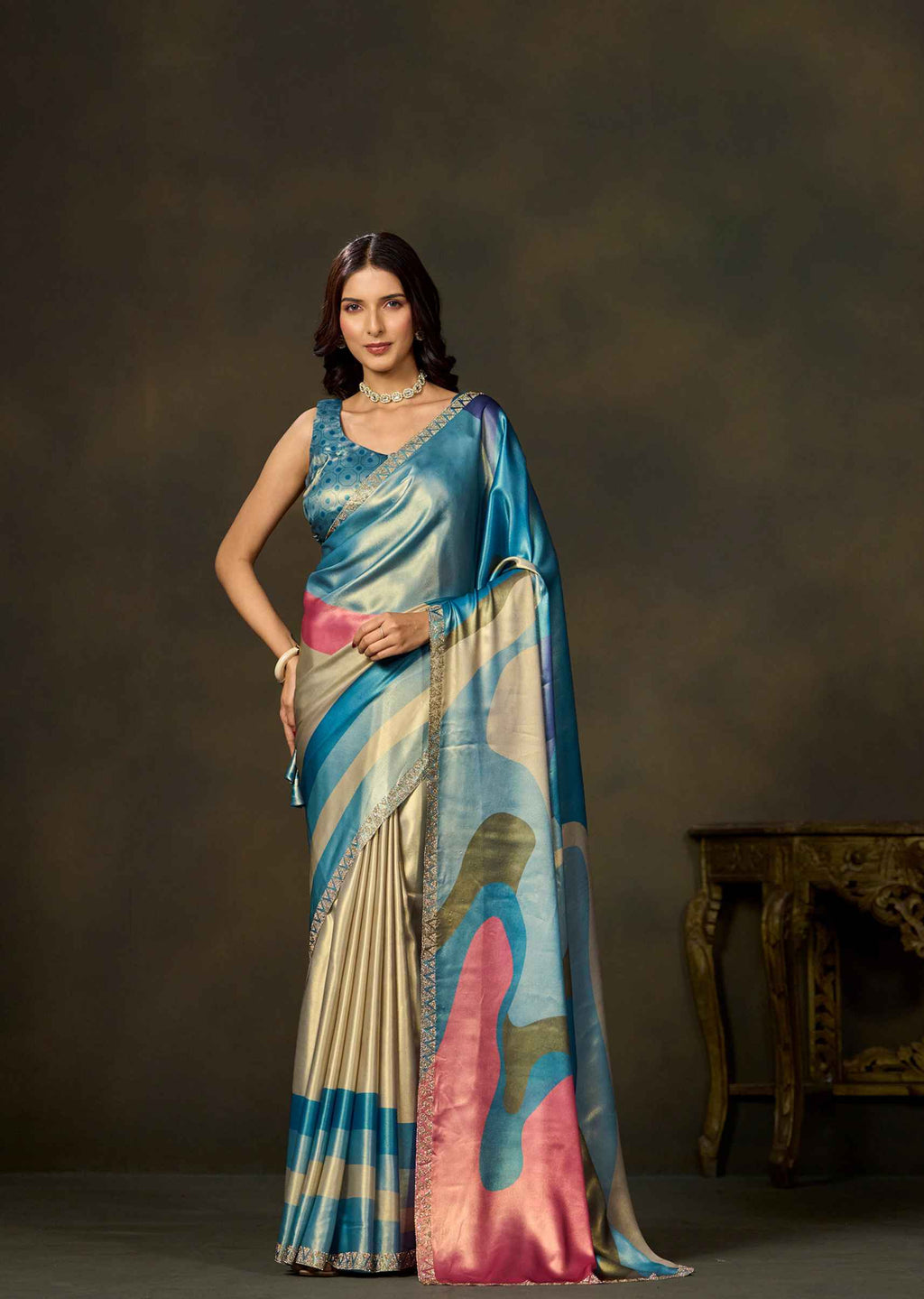 A woman models a traditional Indian saree with a blue, pink, and beige abstract pattern against a dark brown studio backdrop.