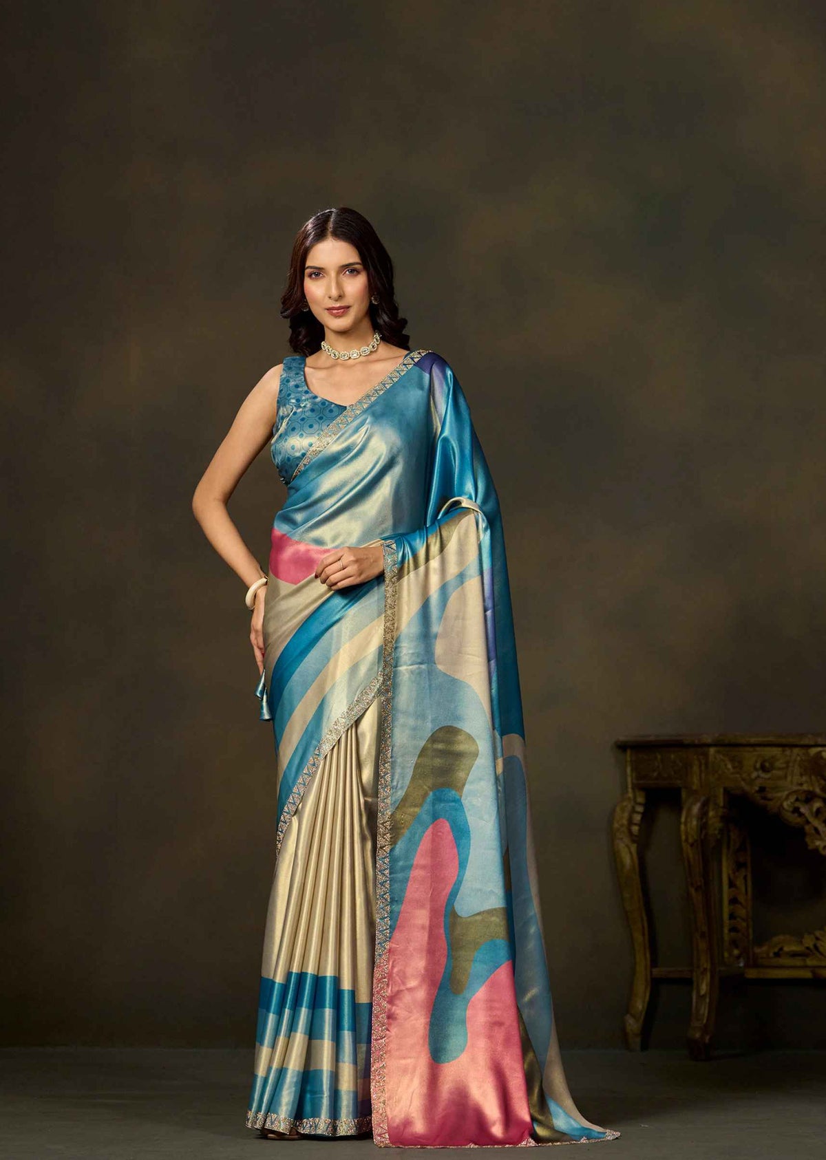 A woman models a traditional Indian saree with a blue, pink, and beige abstract pattern against a dark brown studio backdrop.