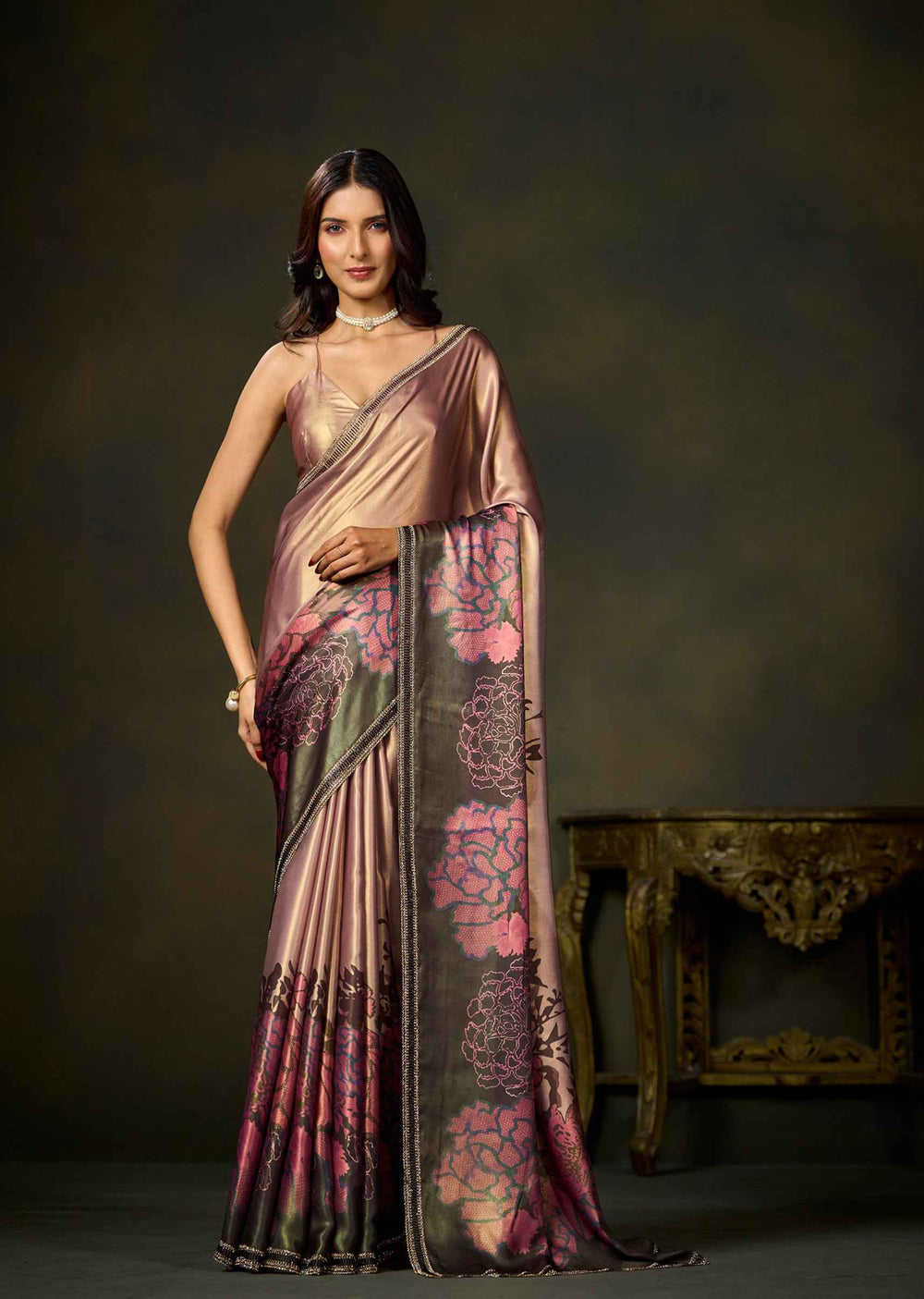 A woman models a metallic rose gold saree with pink floral patterns and a dark border, standing in front of a dark brown studio background with an ornate table.