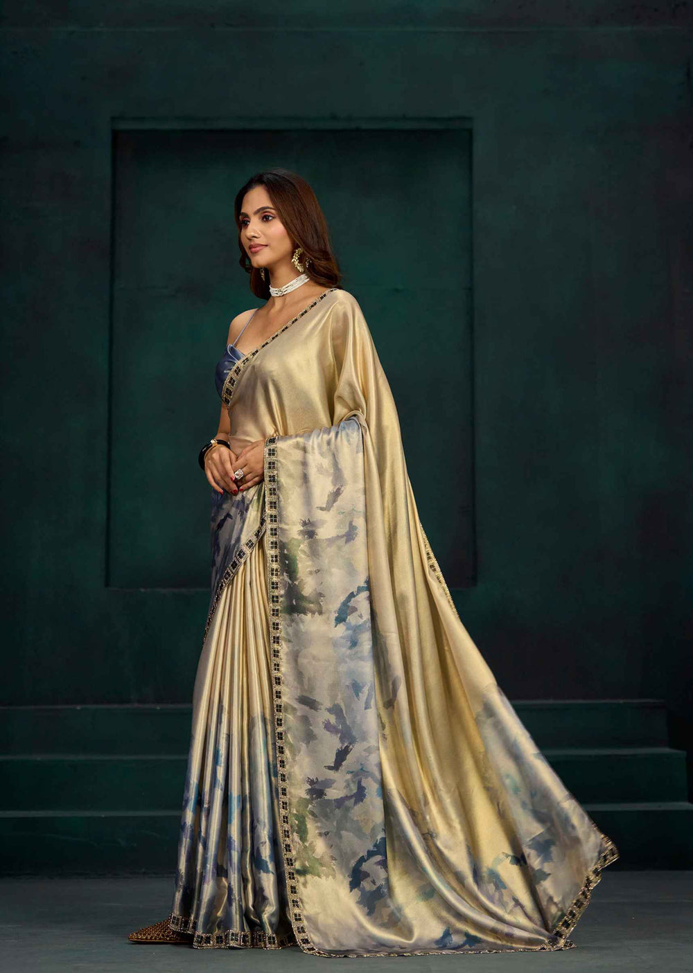 A woman models a luxurious champagne-gold saree with a blue and green floral print border and a matching embroidered blouse, set against a dark teal studio backdrop.