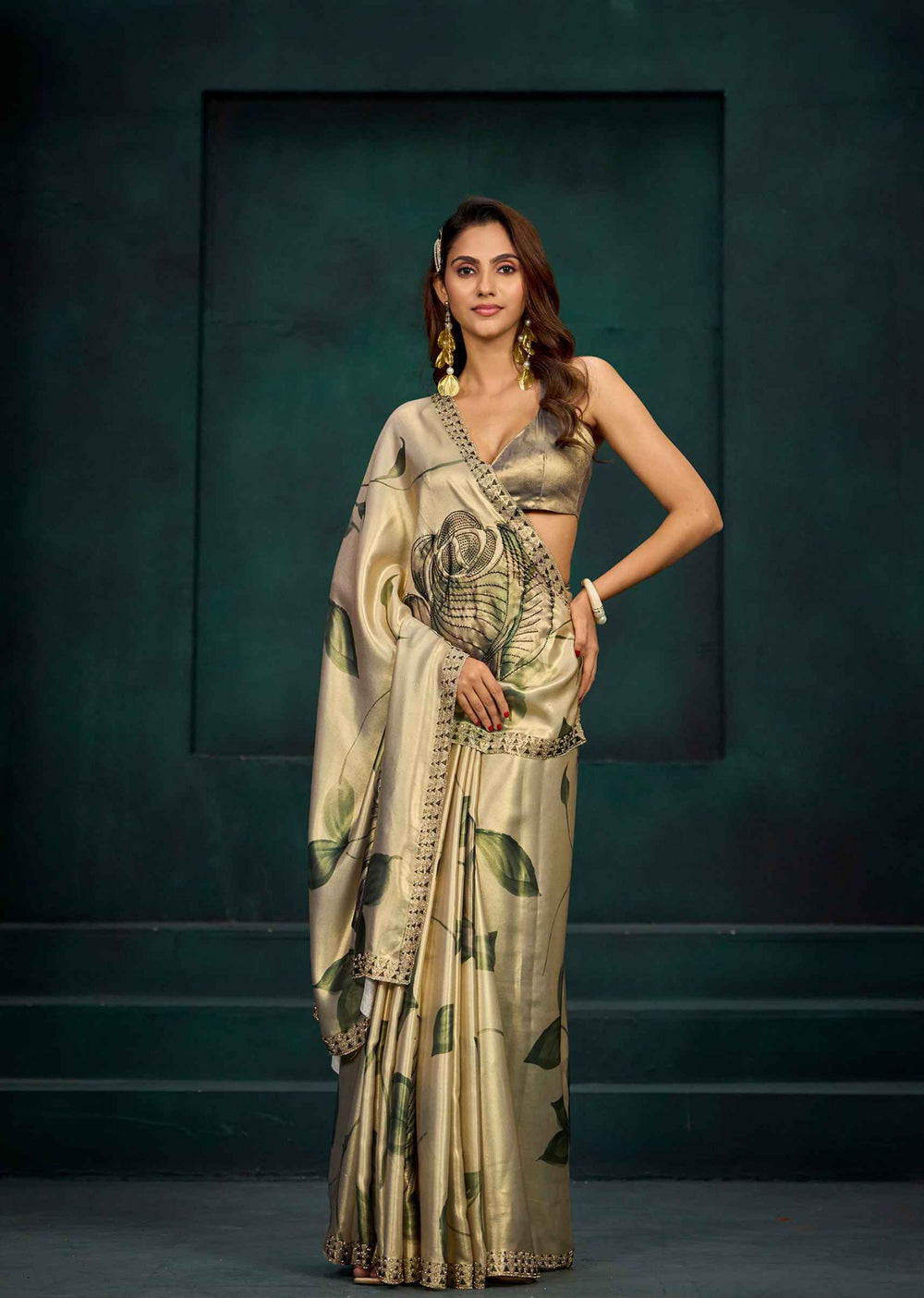 A woman models a metallic gold saree with a green leaf pattern and a matching gold blouse against a dark green studio backdrop.