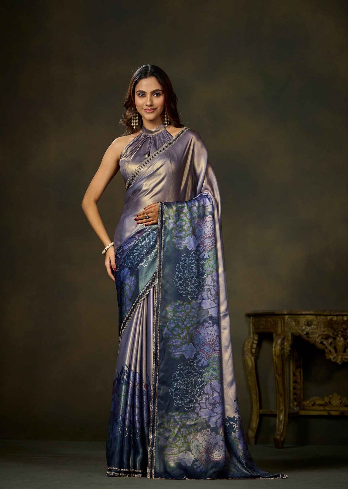 A woman modeling a draped satin saree with a metallic lavender body and a blue floral patterned pallu, posed against a dark brown studio backdrop.