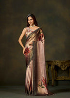 A woman models a draped satin saree with a metallic rose gold base, featuring red floral prints and a black patterned border, set against a dark brown studio background with a decorative table.