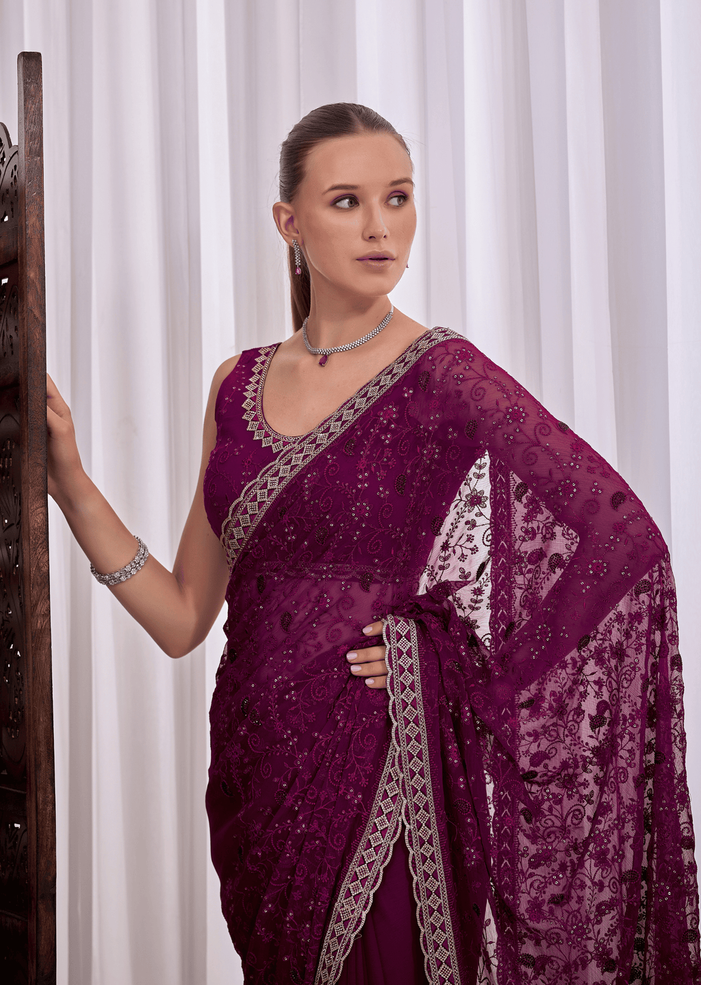 A woman wearing a deep purple embroidered saree with a matching blouse, standing against a white curtain background.