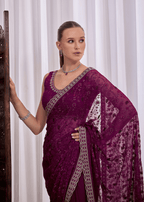 A woman wearing a deep purple embroidered saree with a matching blouse, standing against a white curtain background.