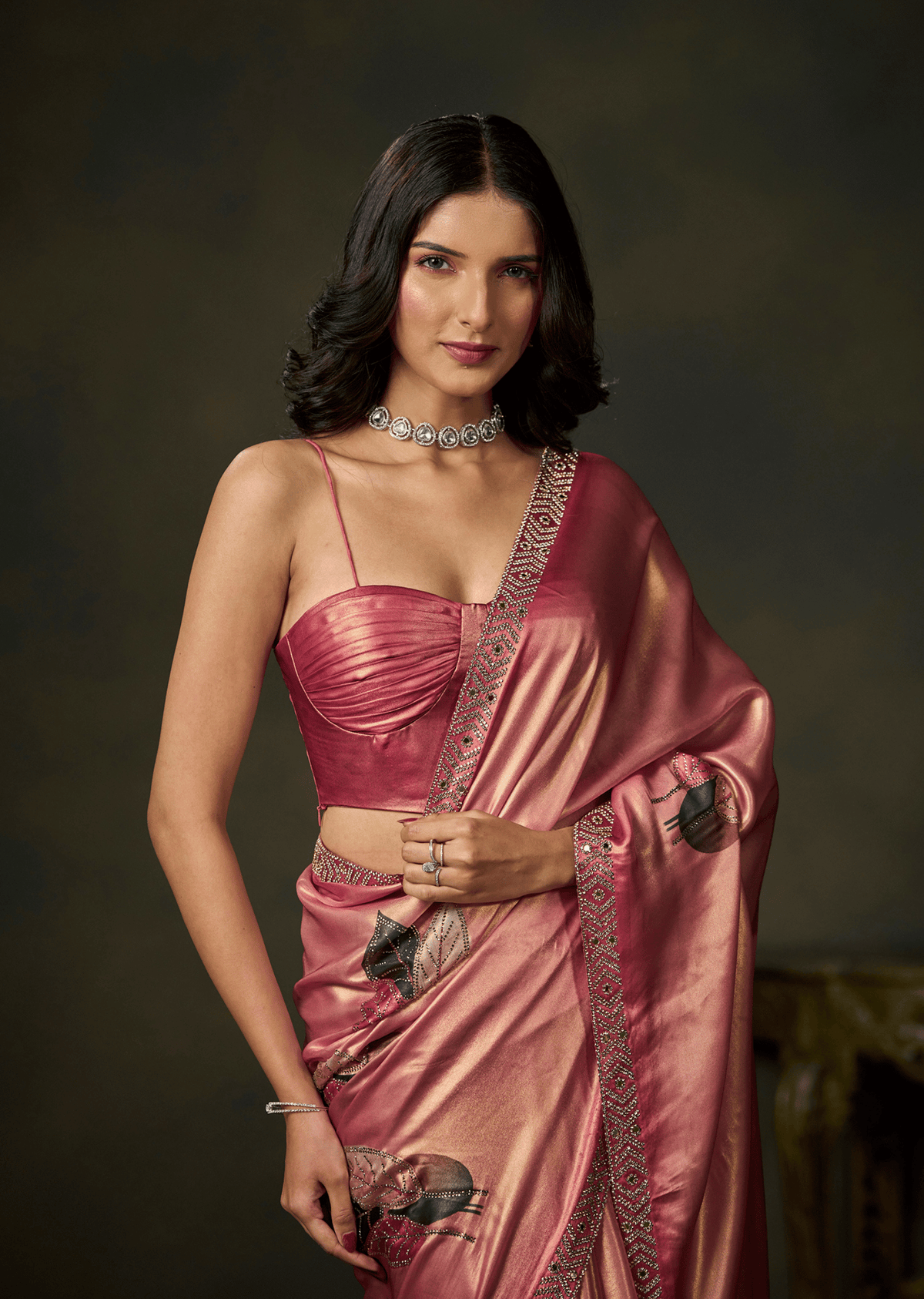 A woman modeling a metallic pink saree with a matching pink blouse, featuring intricate silver embroidery and a silver choker necklace, set against a dark brown studio background.