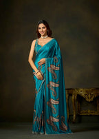 A woman modeling a teal blue saree with brown paisley patterns and gold zari detailing, posed against a dark brown studio backdrop with an ornate table in the background.