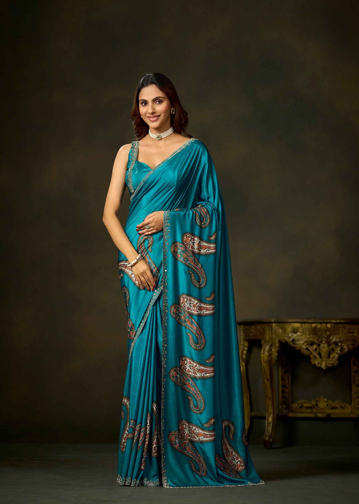 A woman modeling a teal blue saree with brown paisley patterns and gold zari detailing, posed against a dark brown studio backdrop with an ornate table in the background.
