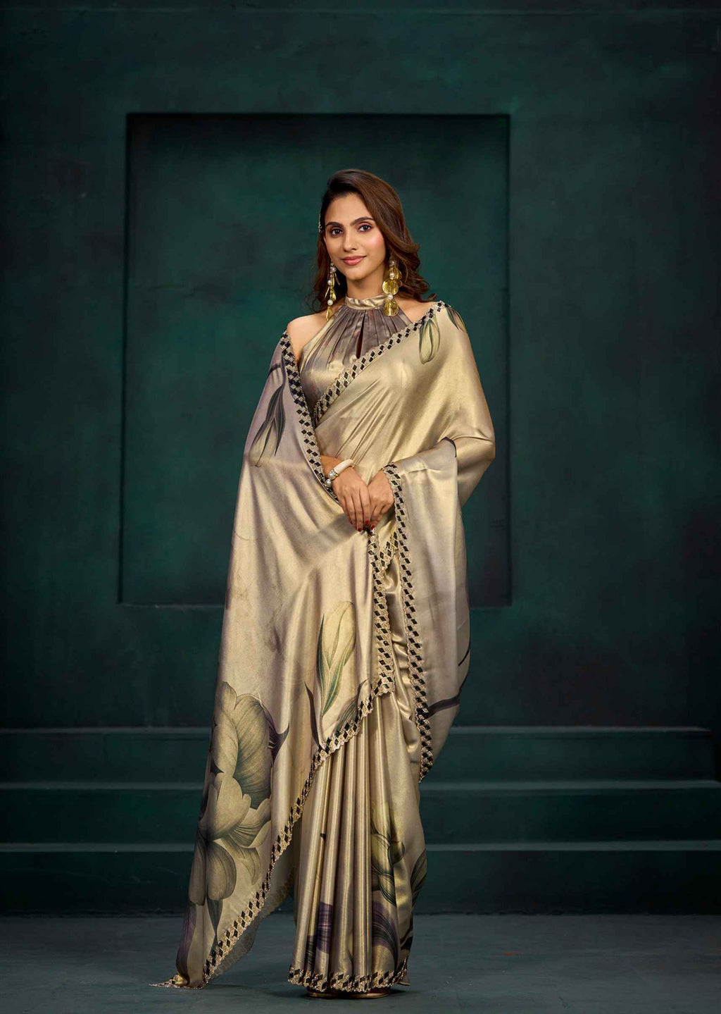 A woman models a draped satin saree with a metallic champagne-gold base, featuring a floral print in muted green and brown tones, and a black-and-gold patterned border. She is posed against a dark, textured studio backdrop.
