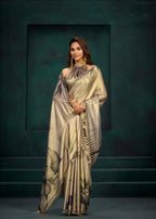 A woman models a draped satin saree with a metallic champagne-gold base, featuring a floral print in muted green and brown tones, and a black-and-gold patterned border. She is posed against a dark, textured studio backdrop.